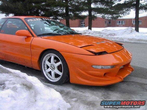 Orange sports car with aftermarket body kit and pop-up headlights, front quarter view in snow