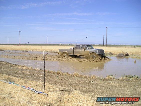 Silver Ford F-Series truck driving through mud, front grille visible