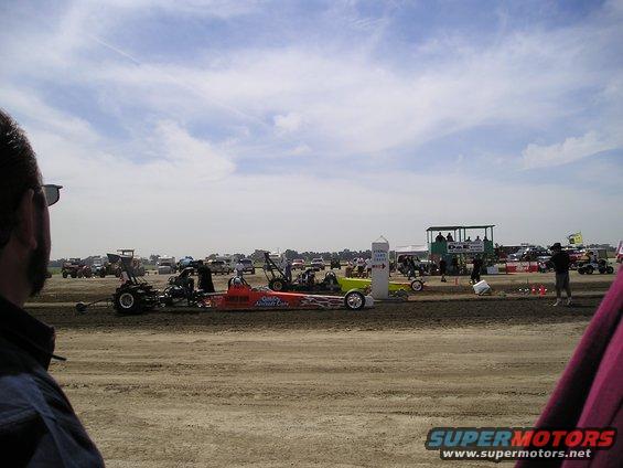 Dragsters lined up on a dirt track with spectators and support vehicles