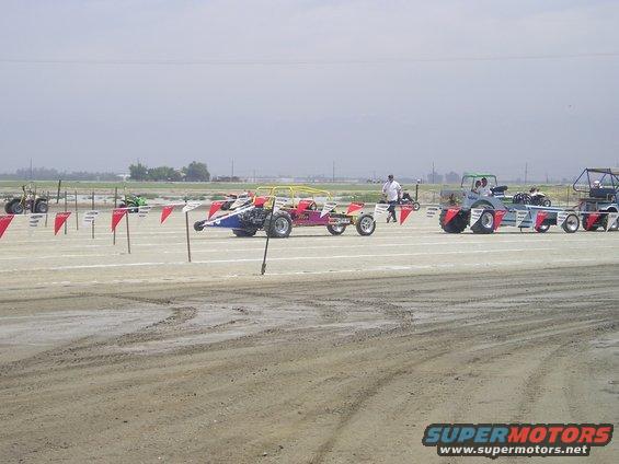 Multiple drag racing vehicles lined up on a dirt track under a cloudy sky