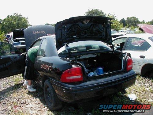 Junkyard scene with a black sedan, trunk open, and a person reaching inside. Other cars are visible in the background