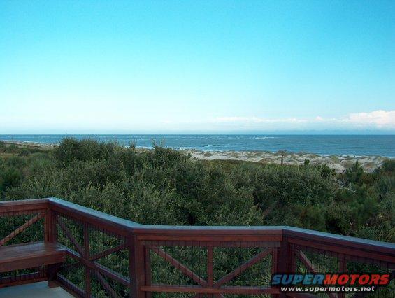 Wooden deck railing overlooks a beach with dunes and ocean under a clear blue sky
