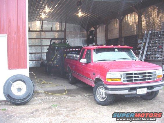 Red Ford F-Series truck, 1995 model year, towing a trailer with a green utility vehicle inside, parked in a barn