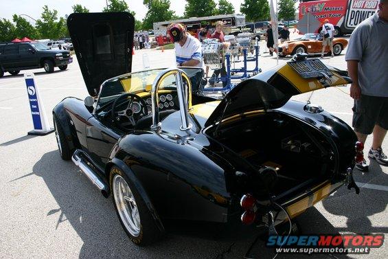 Black AC Cobra replica with yellow stripes, hood and trunk open. Some people and other vehicles in the background