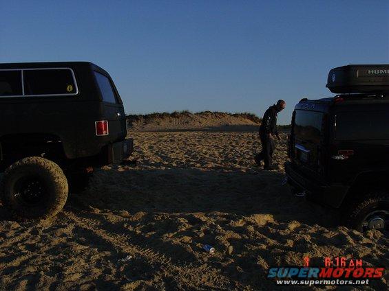 Two black SUVs on sand, one with a roof box. A person walks between them