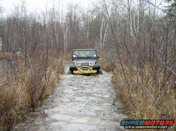 Black Jeep Wrangler with yellow bumper driving on a muddy, icy trail through bare trees