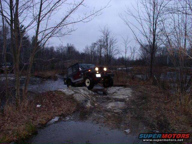 Jeep Wrangler driving through shallow water and over rocks on a trail. Another vehicle is visible in the background