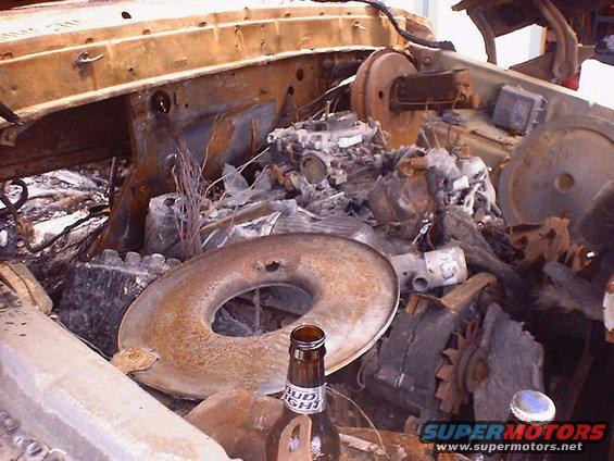 Burnt engine bay with carburetor and air cleaner visible, Bud Light bottle in foreground