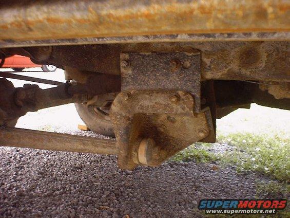 Close-up of heavily rusted vehicle frame with suspension parts visible. Gravel on the ground