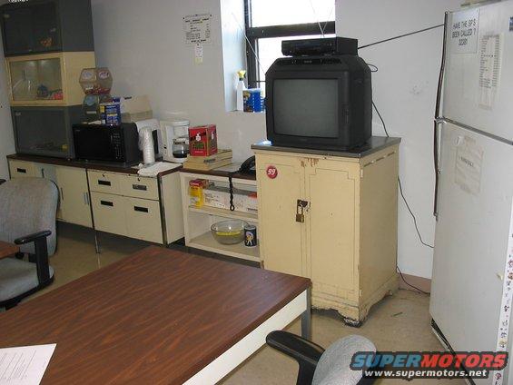 Office breakroom scene with a television on a cabinet and a refrigerator