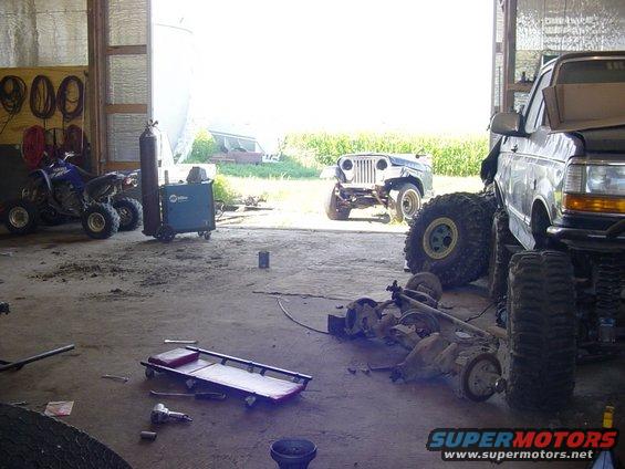 Garage interior with a lifted Ford truck, a disassembled Jeep, an ATV, and various automotive parts scattered around