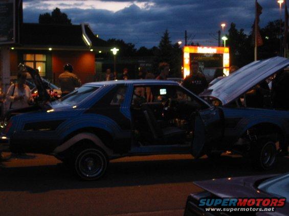 Customized blue coupe with doors and hood open, parked at night. Another car is partially visible in the foreground