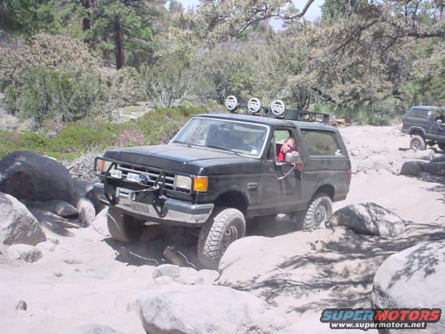 Black Ford Bronco navigating a rocky, sandy trail. Another Bronco is visible behind it