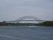 1990 Ford Bronco - A large steel arch bridge spans a wide waterway under a cloudy sky, with a speed boat and sailboats visible.