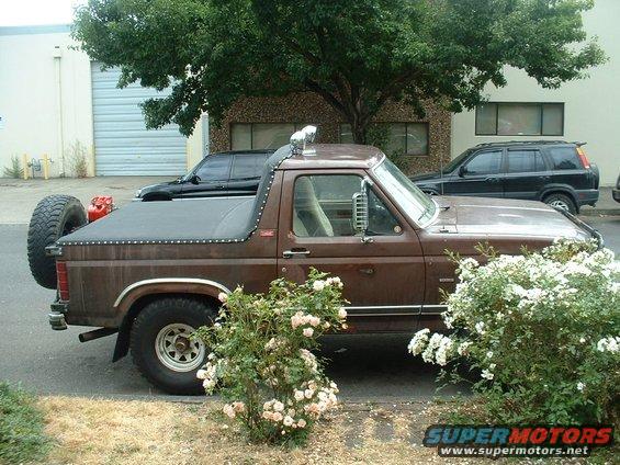 Brown pickup truck with a spare tire mounted on the rear and a black tonneau cover. A red gas can is visible behind the cab. Another black SUV and a black sedan are parked behind it