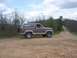 1984 Ford Bronco - A two-tone brown Ford Bronco is parked on a gravel road surrounded by trees under a cloudy sky.