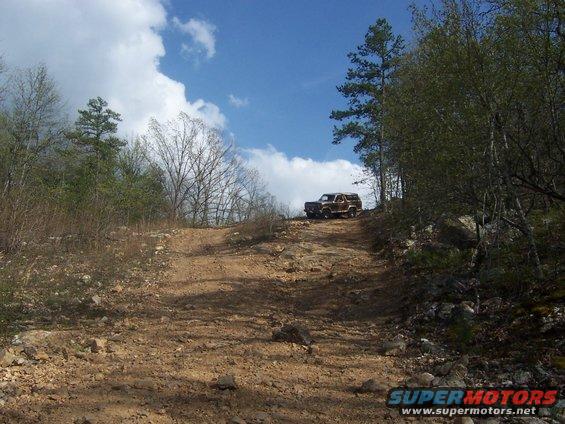 Brown SUV on a rocky dirt path uphill, trees and blue sky with clouds