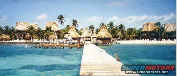 Tropical resort with thatched-roof bungalows on a white sand beach, viewed from a wooden pier over turquoise water