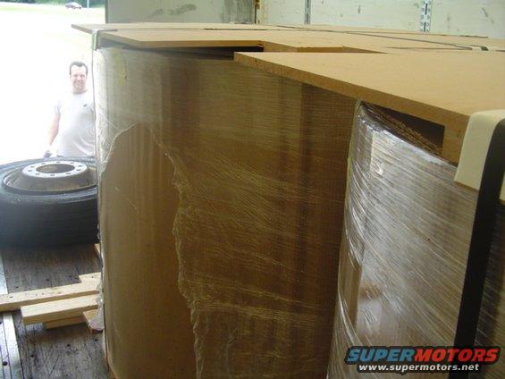 Large cylindrical objects wrapped in plastic and cardboard, with a man and a tire visible in the background