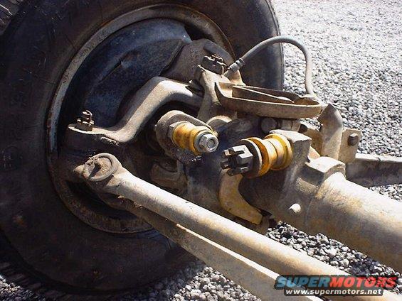Close-up of a vehicle's front axle and suspension with a tire visible. Rusty metal parts and yellow bushings are prominent