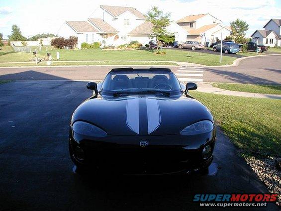 Black convertible sports car with dual white racing stripes parked in a driveway. Houses and other cars visible in the background