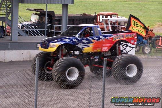 Monster truck with stars and stripes paint scheme, driving past a fence. Other trucks and tractors are in the background