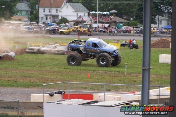 Blue monster truck with "Broker" on the side, driving on a grassy field with other cars and spectators in the background