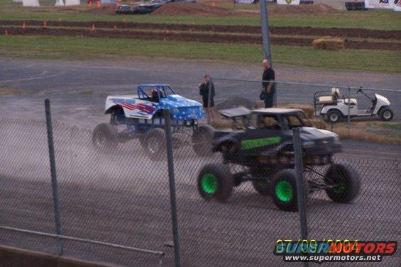 Two monster trucks race on a dirt track, one with patriotic paint, the other black with green wheels. A golf cart is parked nearby