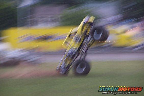Yellow monster truck on its rear wheels with large tires, blurred motion. Supermotors.net logo visible