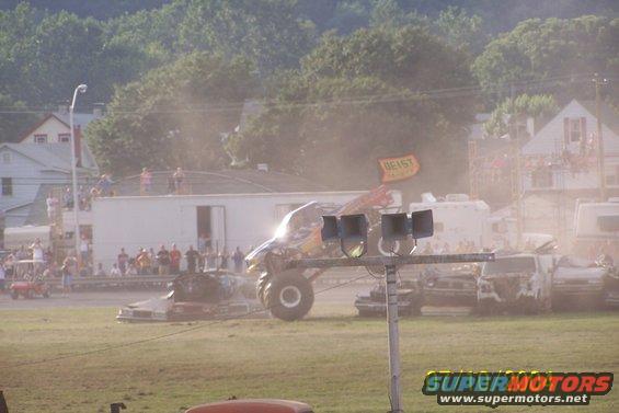 Monster truck crushing cars in a demolition derby event. A flag with 'GEIST' is visible