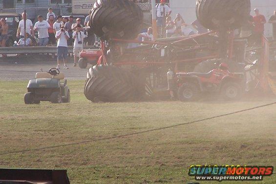 Monster truck on its side with an ATV nearby and a golf cart in the foreground. Spectators watch from a raised platform