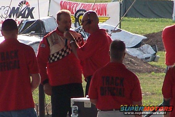 People in red 'TRACK OFFICIAL' shirts stand near a damaged vehicle at an outdoor event