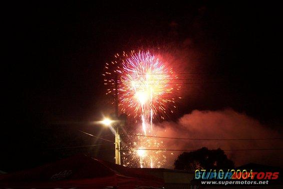 Fireworks explode in the night sky over a dark landscape with a street light and some smoke