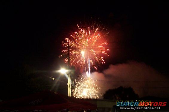 Fireworks explode in the night sky over a streetlamp and silhouetted buildings