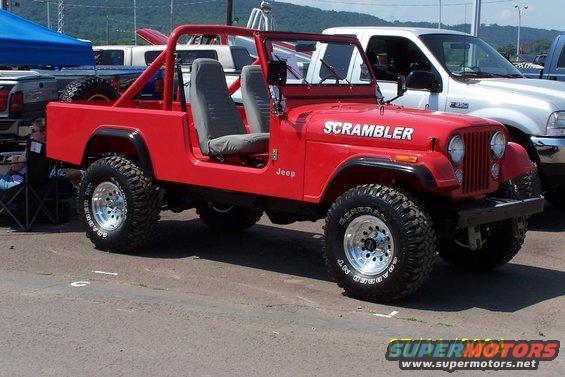 Red Jeep CJ-8 Scrambler with roll cage and aftermarket wheels. White pickup truck is parked next to it