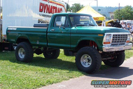 Green pickup truck with large tires and lift kit parked on grass, with a trailer and tents in the background