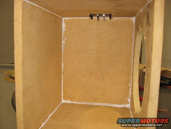 Interior of an unfinished wooden speaker box with sealant visible on the edges. Some writing is scrawled on the back panel