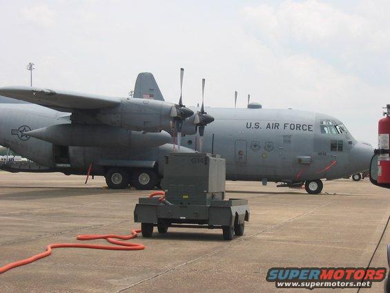 Gray U.S. Air Force C-130 Hercules aircraft on tarmac with a ground power unit and orange hose