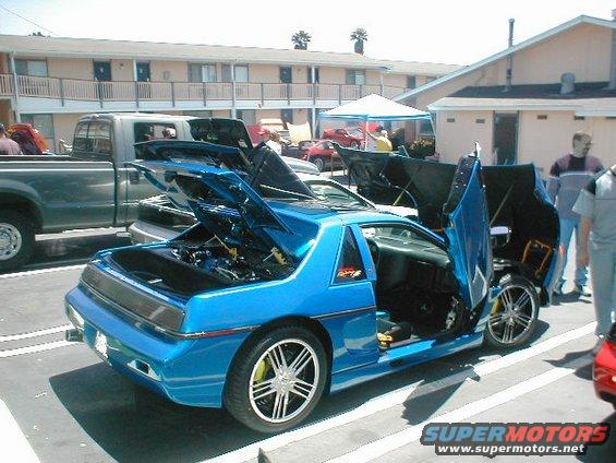 Blue 1986 Pontiac Fiero with gullwing doors and hood open, custom wheels, parked next to a pickup truck