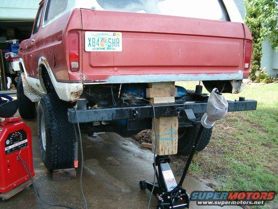 Red and white 1978 Ford Bronco rear end raised on blocks and jack, with a welding machine nearby