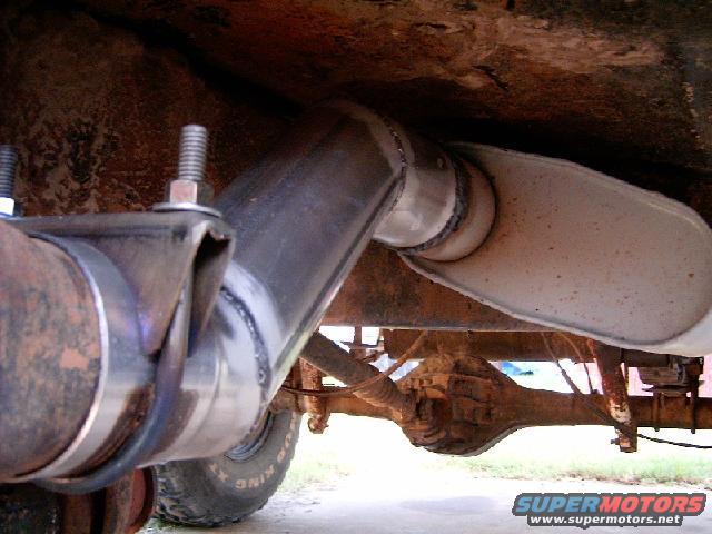 Exhaust pipe and muffler assembly attached to a rusty rear axle. Visible text: SUPERMOTORS.NET