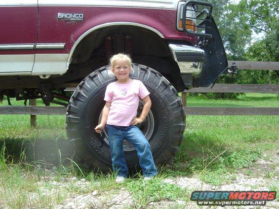 Maroon and white Ford Bronco with oversized tires, a young girl stands in front of the front passenger tire