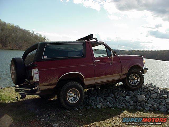Maroon Ford Bronco parked on rocks by a lake. Spare tire on the back