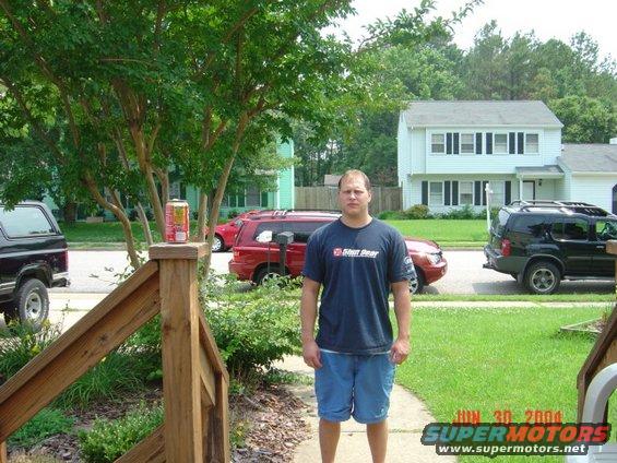 Man in blue shirt and shorts standing on sidewalk with vehicles and houses behind him