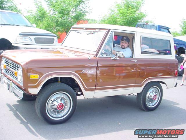Brown 1979 Ford Bronco with white top, driver visible. Another truck is parked to the left