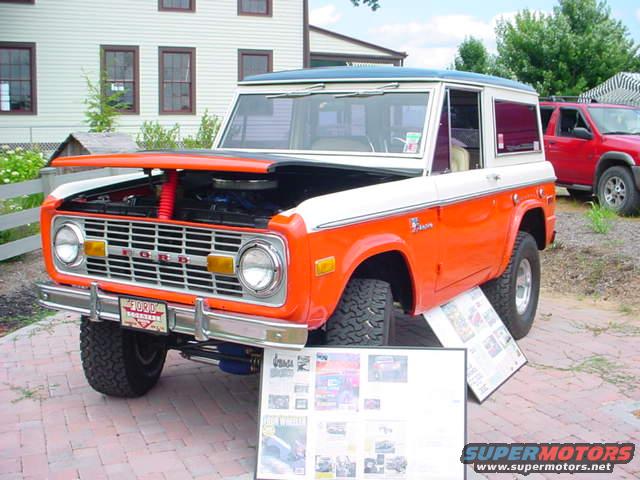 1979 Ford Bronco with hood open, parked on brick pavers. Another red SUV is visible behind it