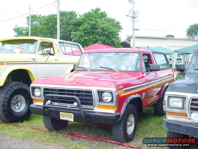 Red 1978 Ford Bronco with yellow and orange side stripes, parked next to a yellow Bronco and a dark vehicle