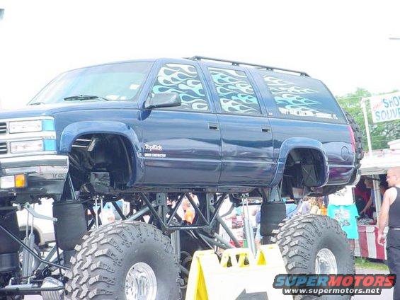 Blue monster truck with flame decals on windows, lifted high on massive tires