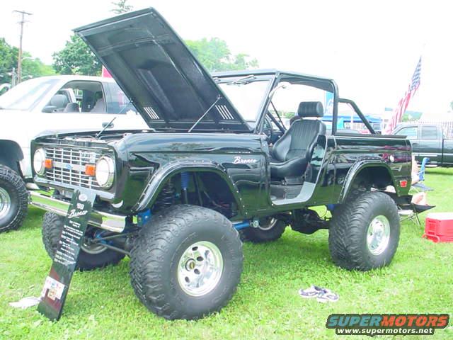 Black 1979 Ford Bronco with hood open, lifted suspension, and aftermarket wheels. A white truck is parked behind it