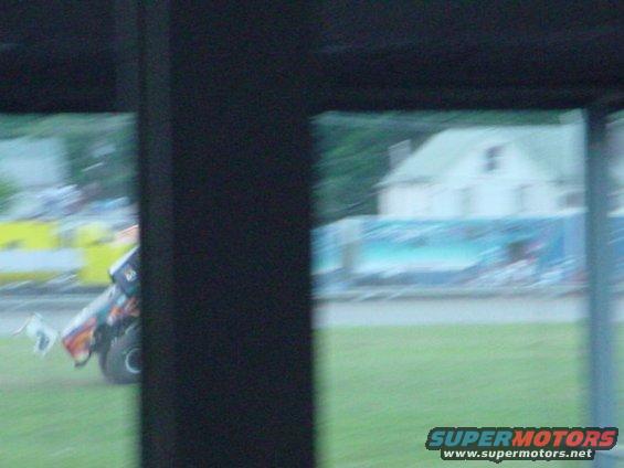 Monster truck with colorful graphics on its side, seen from the side, with a building and a fence in the background